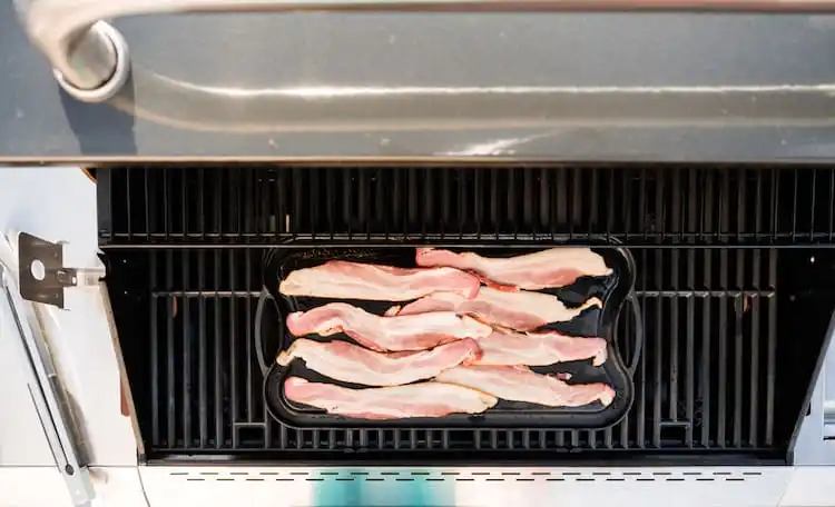 Frying bacon on cast iron griddle in outdoor gas grill.